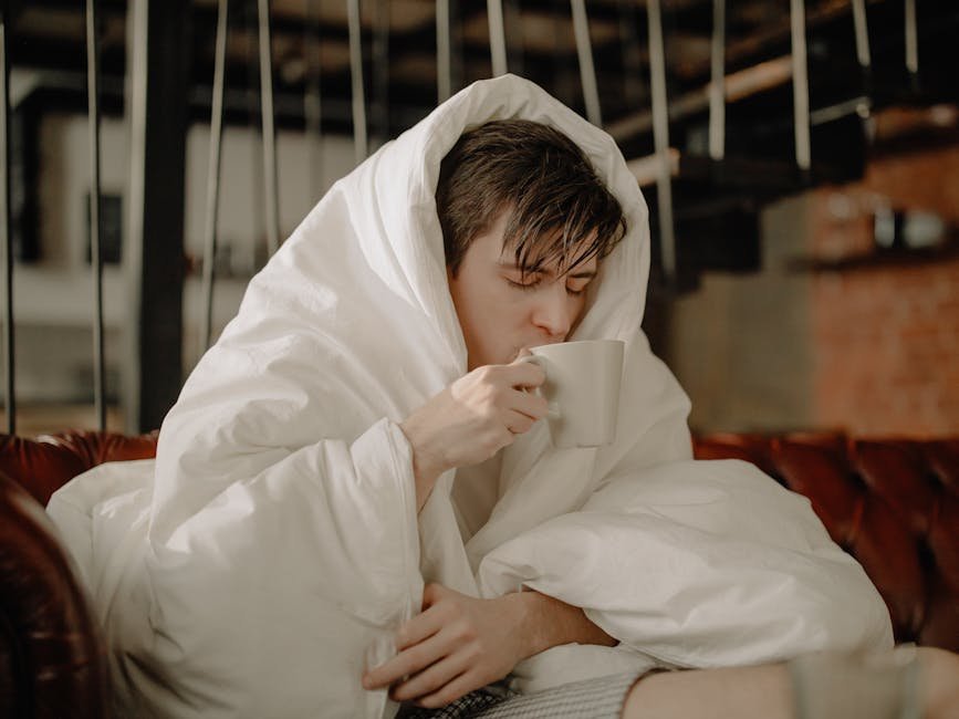 Young man wrapped in blanket sipping hot beverage at home, embracing comfort and relaxation.